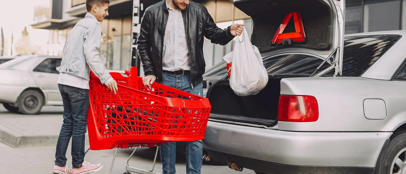 Férfi és egy kisfiú pakol bevásárlószatyrokat egy autó csomagtartójába egy áruház parkolójában, mellettük piros bevásárlókocsi